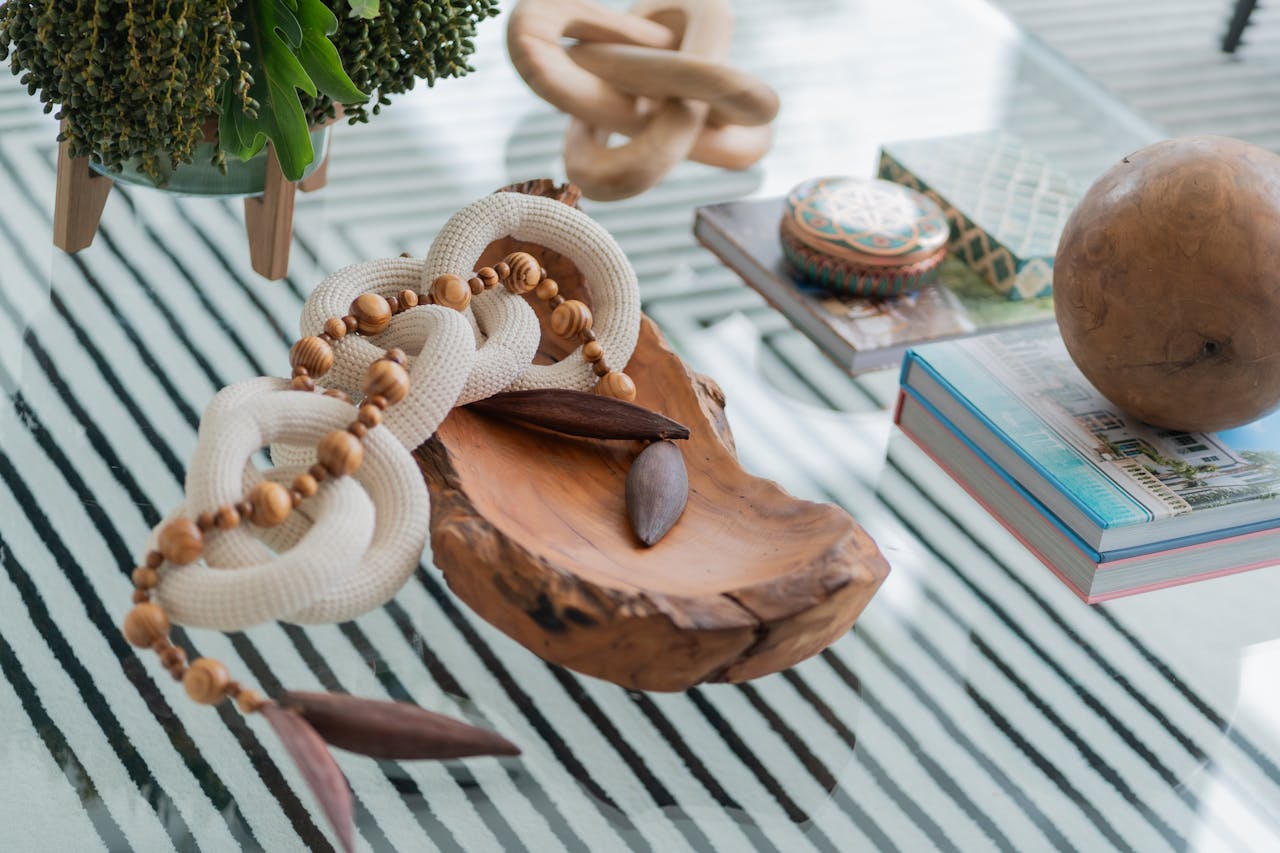 A stylish arrangement of decorative objects on a glass table, featuring a wooden bowl, beads, and greenery.