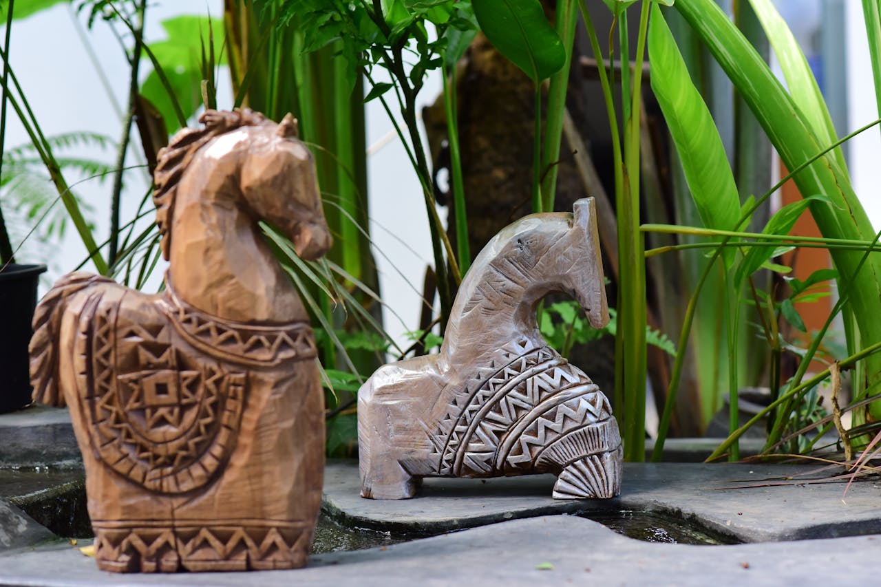 Two handcrafted wooden horse sculptures displaying intricate patterns set amidst lush greenery.